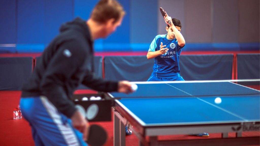 Atleta treinando tênis de mesa em clube, recebendo bolas do treinador e executando golpe ofensivo com foco na técnica.