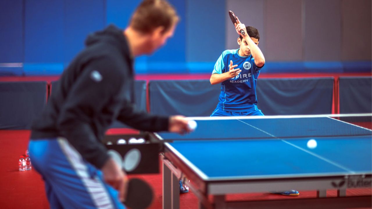 Atleta treinando tênis de mesa em clube, recebendo bolas do treinador e executando golpe ofensivo com foco na técnica.
